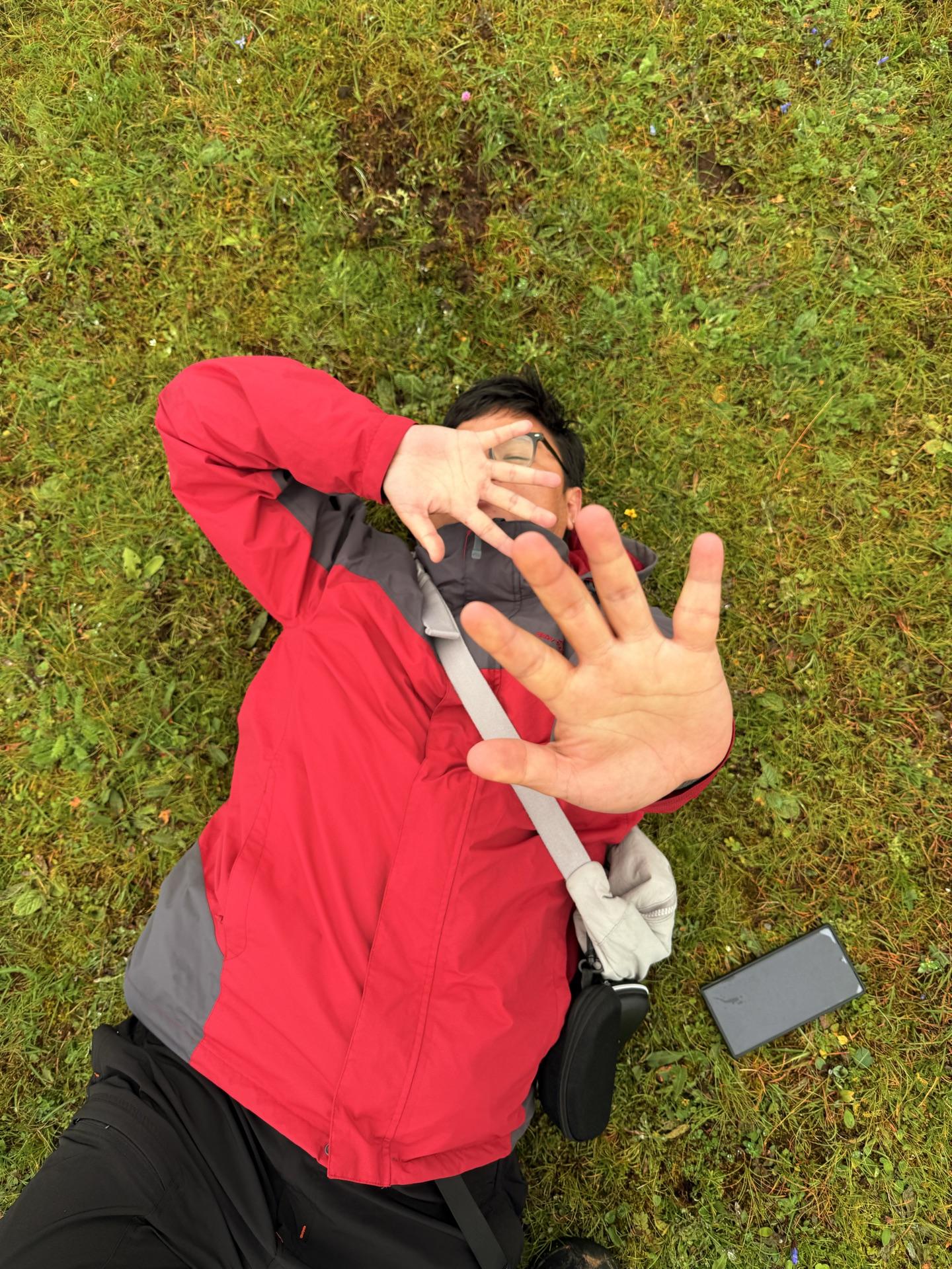 A friend in a red jacket lying on the meadow grass, hand raised to block the camera