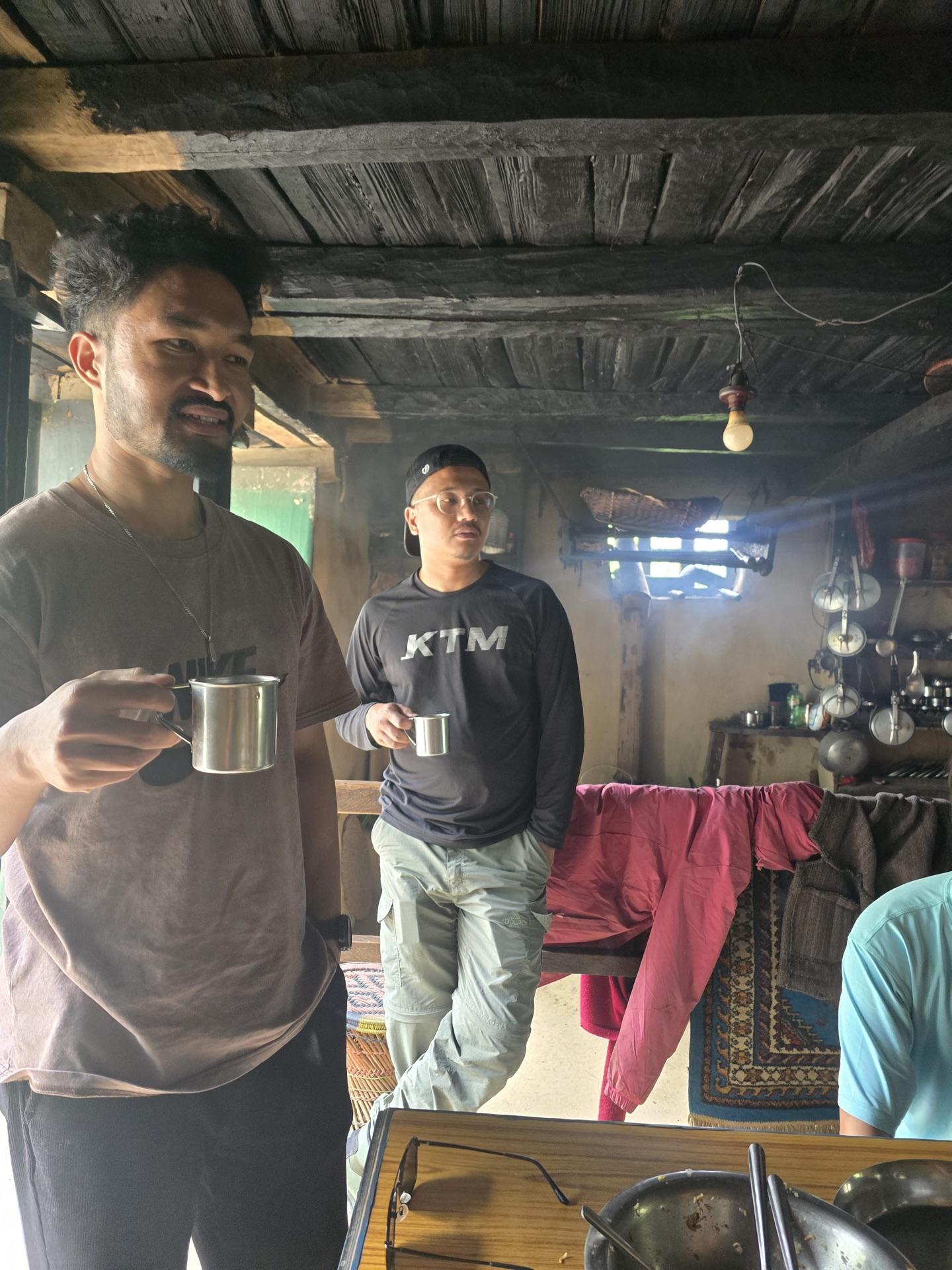 Two friends raising steel cups of tea inside a wooden roadside kitchen