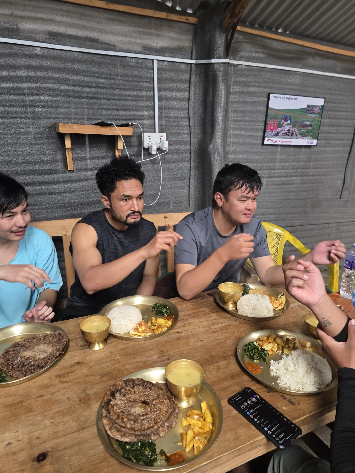 Four friends around a wooden table eating dal bhat from brass plates