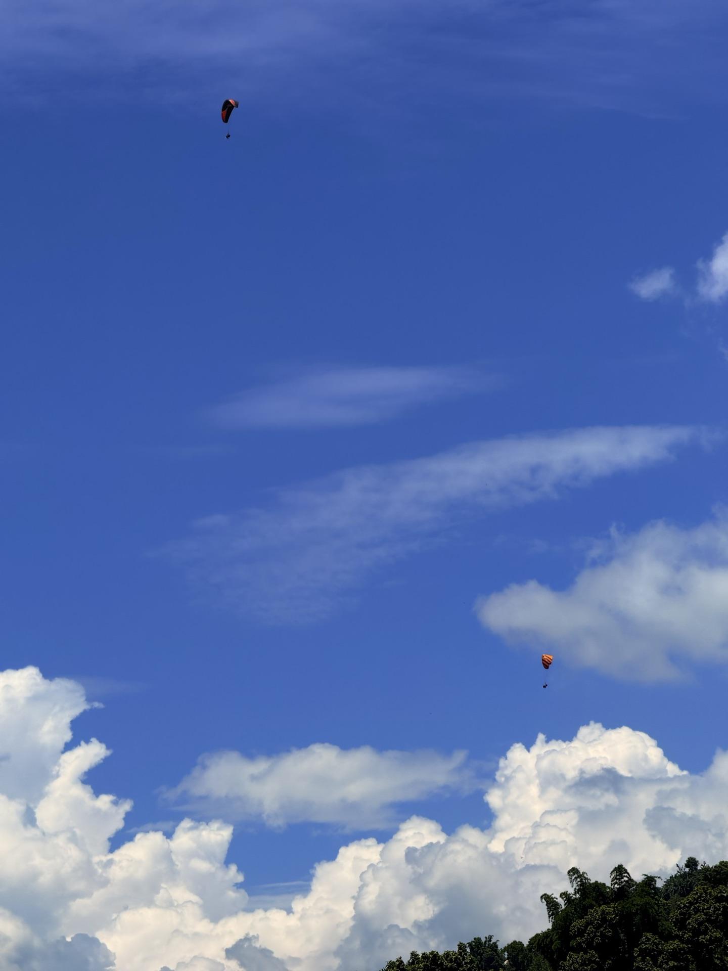 Two tiny paragliders high in a blue sky above white clouds