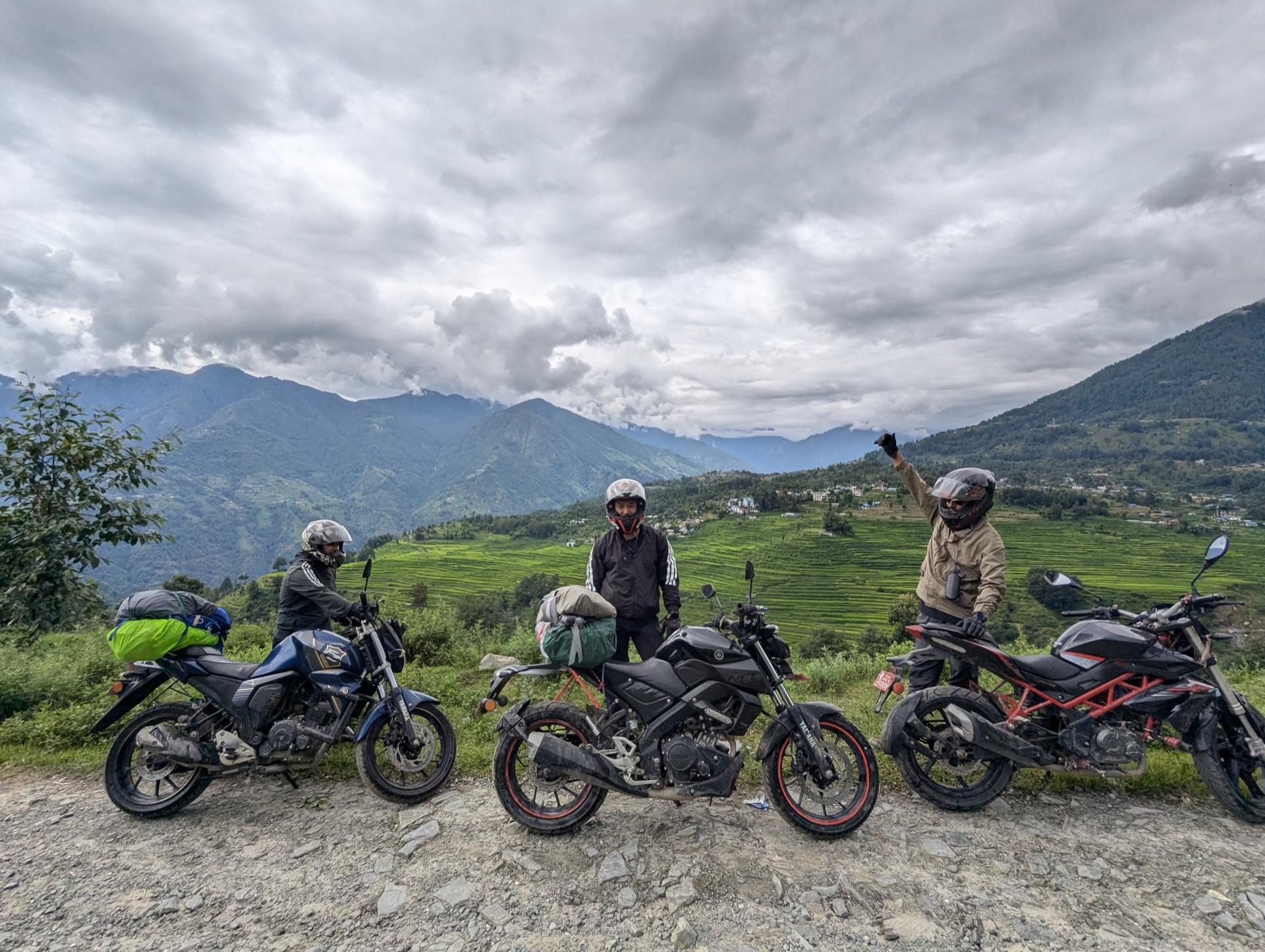 Three loaded motorbikes parked on a gravel bend while the riders rest, terraced hills behind