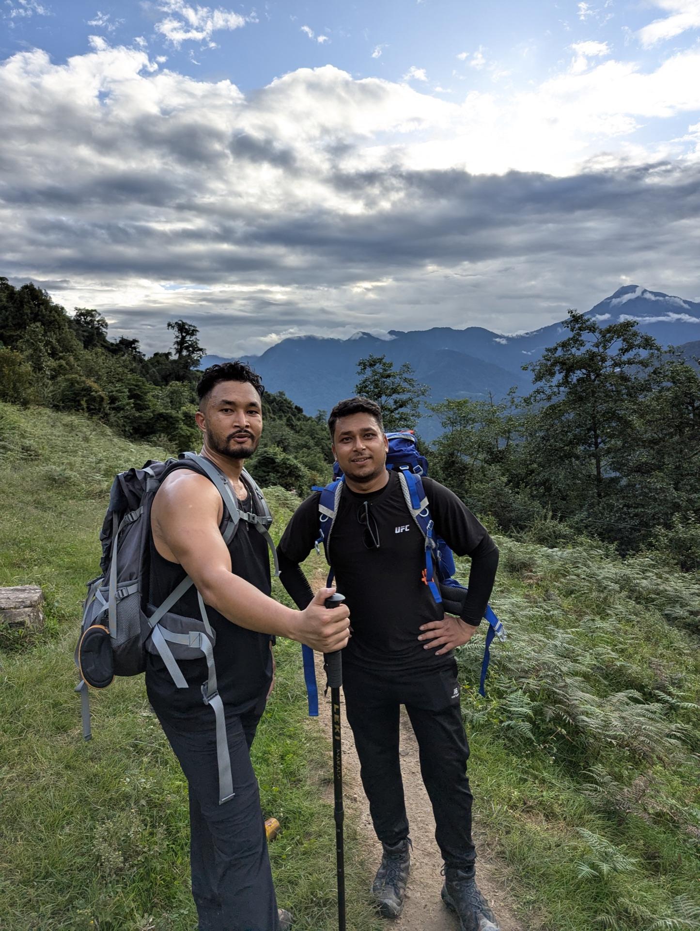 Two friends with backpacks climbing a green hillside trail in evening light